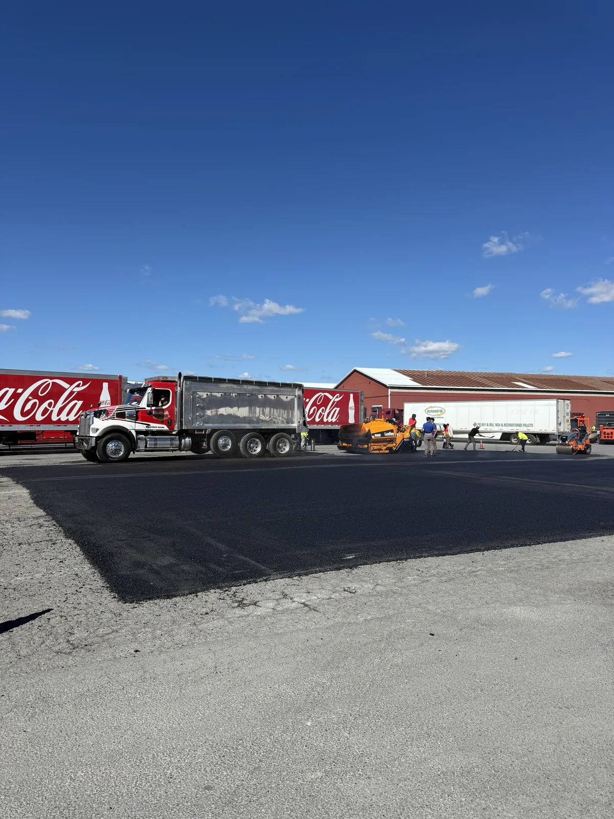 A newly paved asphalt area in a commercial yard with parked trucks beyond.