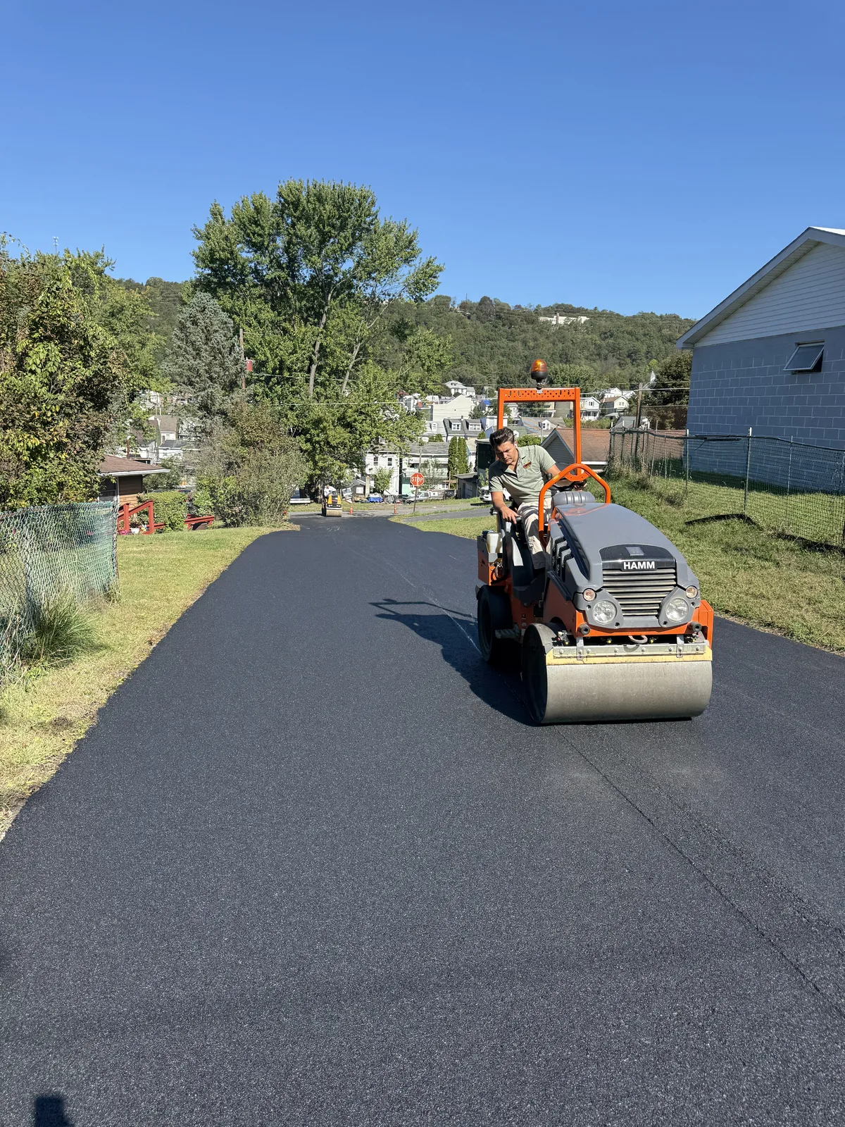 Operator compacting asphalt with a roller on a sloping street between buildings.