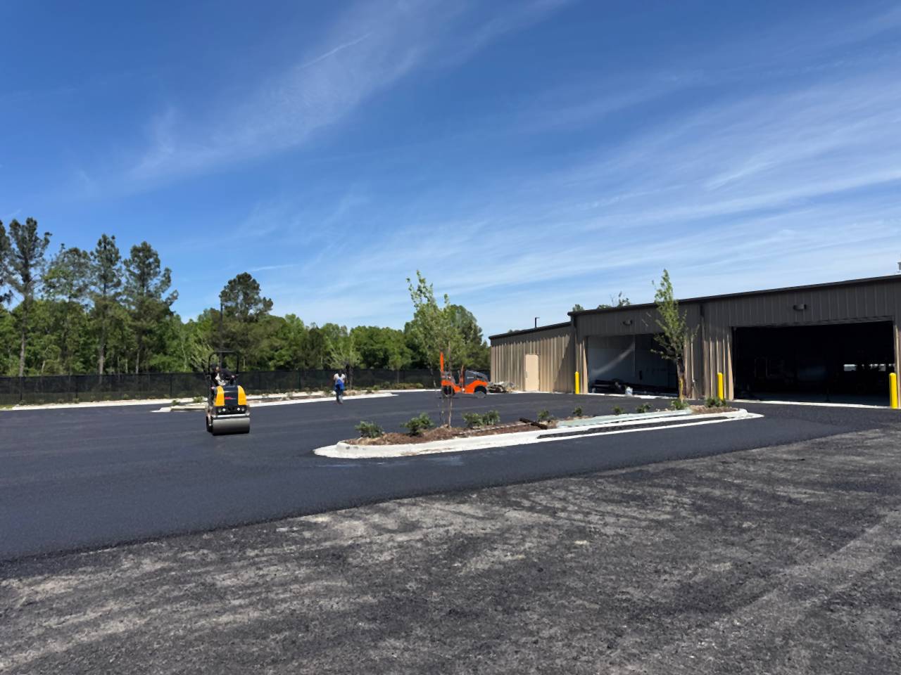Fresh asphalt and curbed islands at the Caliber Collision new construction site in Denham Springs, Louisiana, with a roller beside the building.