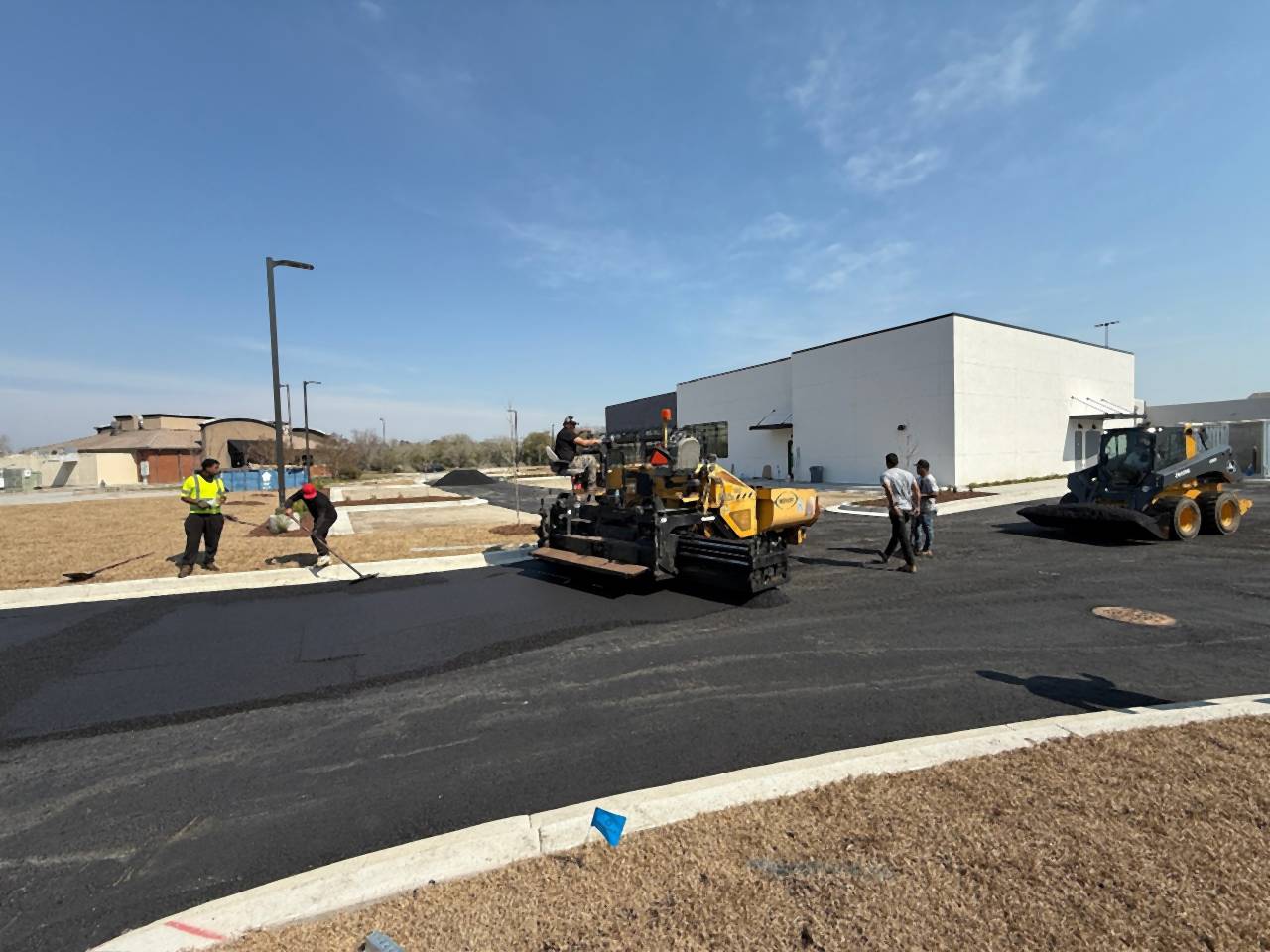 Paver, roller and crew placing asphalt beside new curbs at a commercial building.